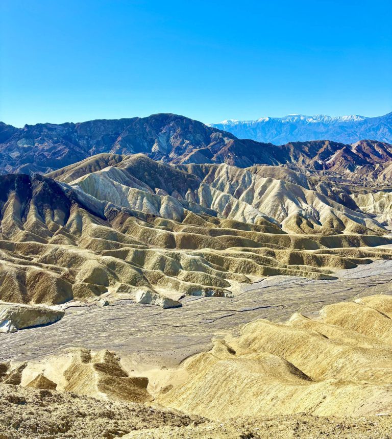 Panoramic view of rugged, multicolored desert badlands with deeply eroded hills and distant snow-capped mountains beneath a clear blue sky near Las Vegas.